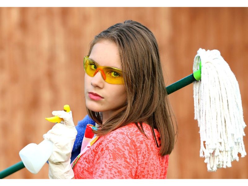 Woman in goggles and apron facing camera holding spray bottle, rag and mop for DIY home cleaning