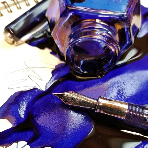 Royal blue ink spilled onto notebook beside fountain pen