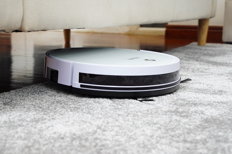 Round robotic vacuum cleaner under couch on white rug in living room with hardwood floors