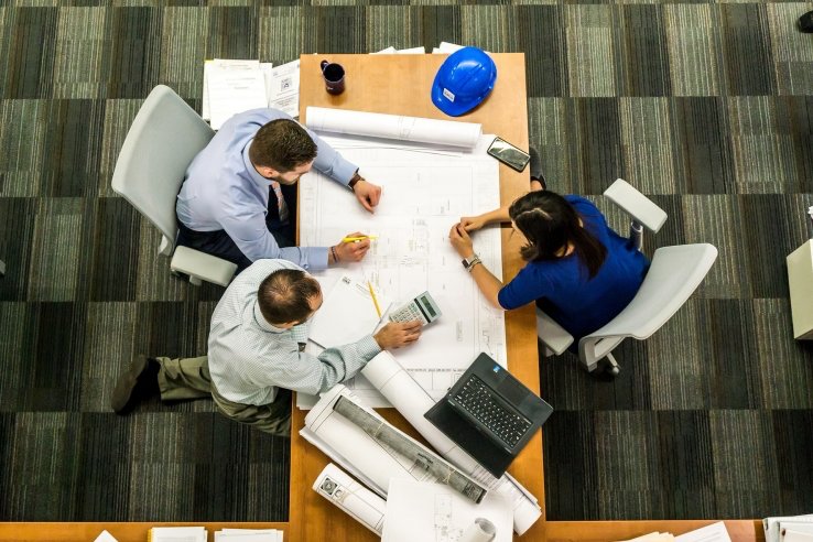 Coworkers in carpeted office seated at table with blueprint and laptop