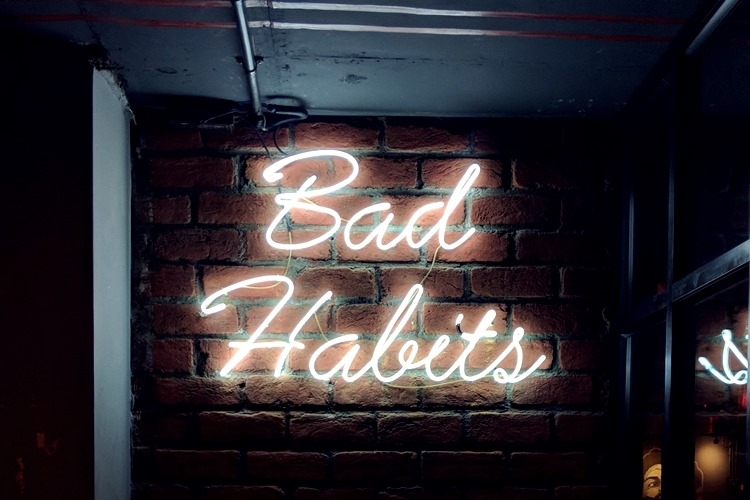 Neon sign with 'Bad Habits' in cursive on indoor brick wall