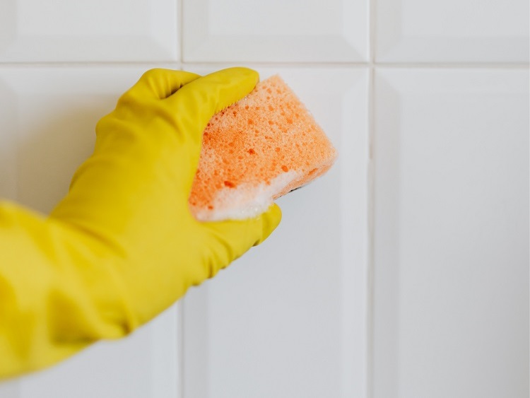 Hand in yellow rubber glove cleaning while tiled wall with beige grout using orange sponge