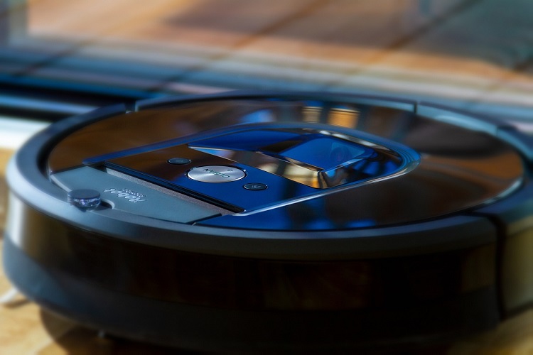 Black robotic vacuum cleaner model on light-colored wooden floor beside glass door