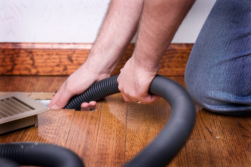Adult kneeling on wooden floors cleaning air ducts with a vacuum through an open air vent